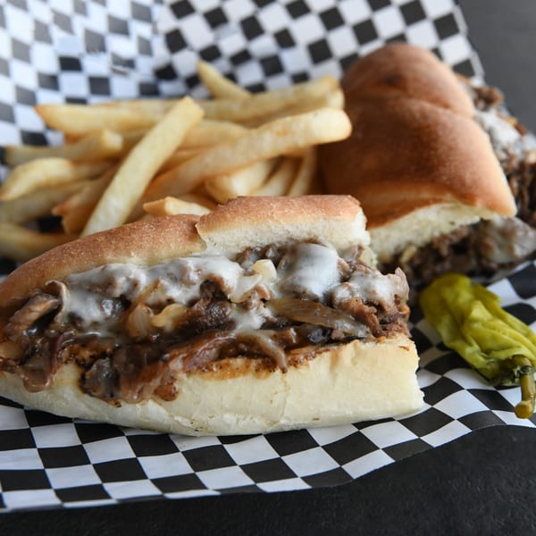 Cheesesteak sandwich with melted cheese, onions, and mushrooms, served with fries and a pepper.