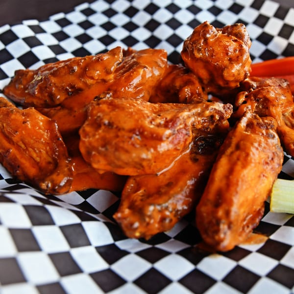 Milano's Pizzeria's buffalo wings, coated in orange sauce, served in a checkered paper basket.