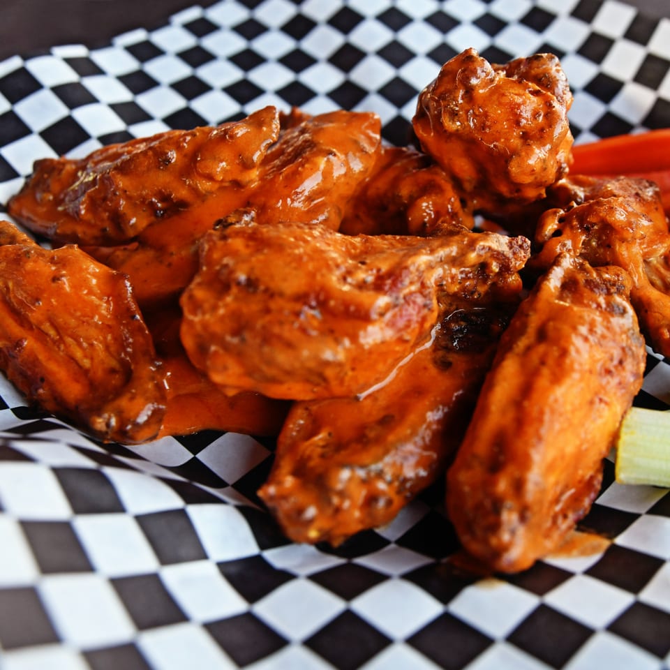 Milano's Pizzeria's buffalo wings, coated in orange sauce, served in a checkered paper basket.