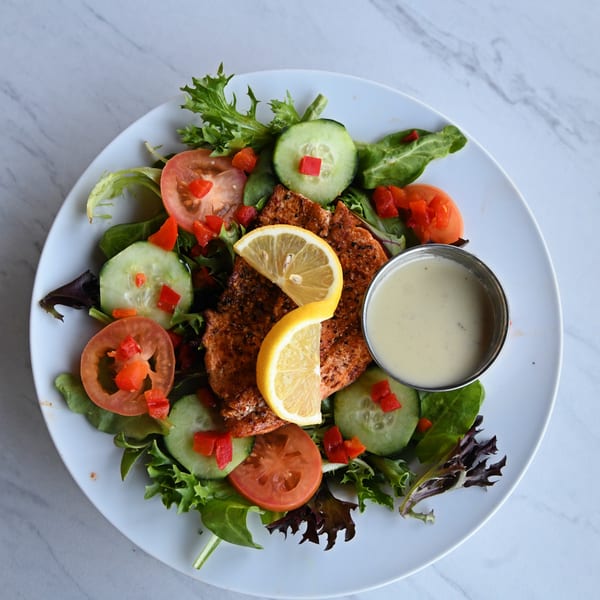 Grilled salmon salad with mixed greens, tomatoes, cucumbers, red peppers, lemon, and dressing.